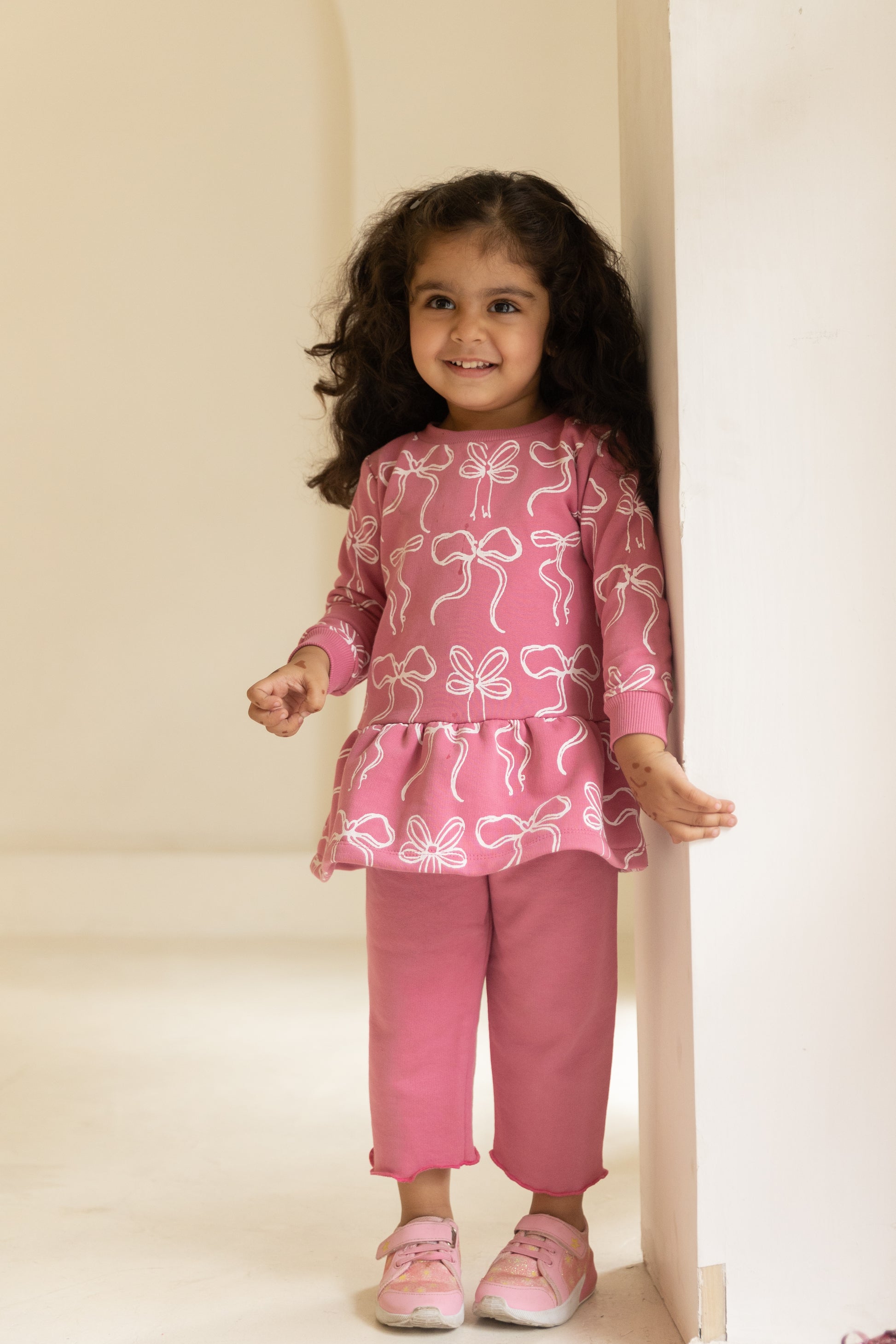 Young girl wearing a pink outfit with butterfly pattern standing against a white wall.