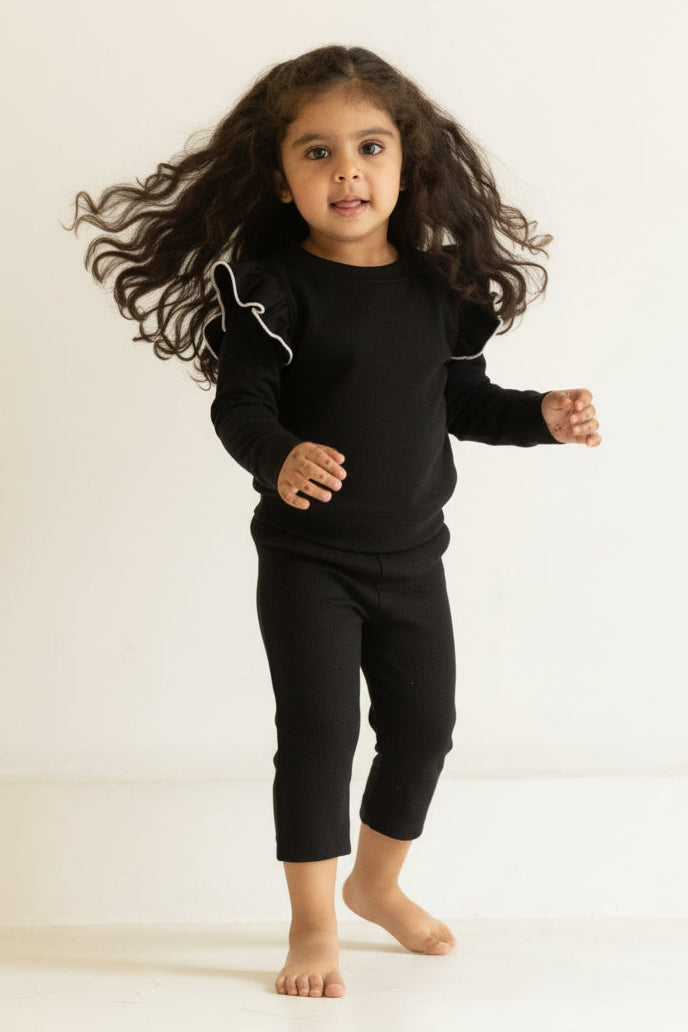 Child wearing a black outfit standing in a minimalistic room with beige walls.