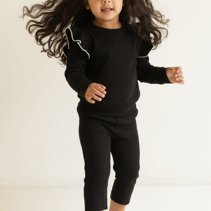 Child wearing a black outfit standing in a minimalistic room with beige walls.