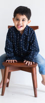 Child sitting on a wooden stool wearing a navy blue shirt with white patterns and light blue jeans.