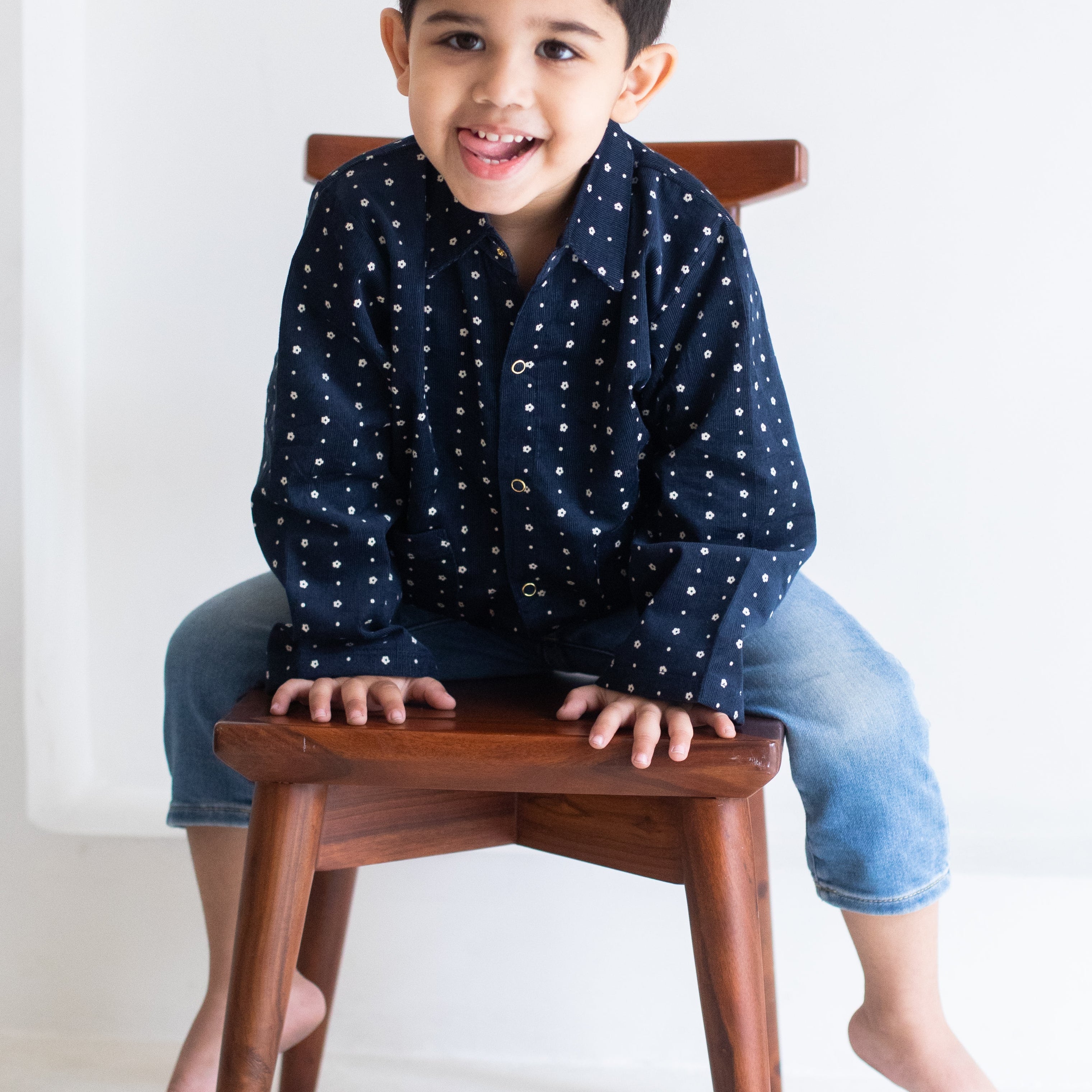 Child sitting on a wooden stool wearing a navy blue shirt with white patterns and light blue jeans.