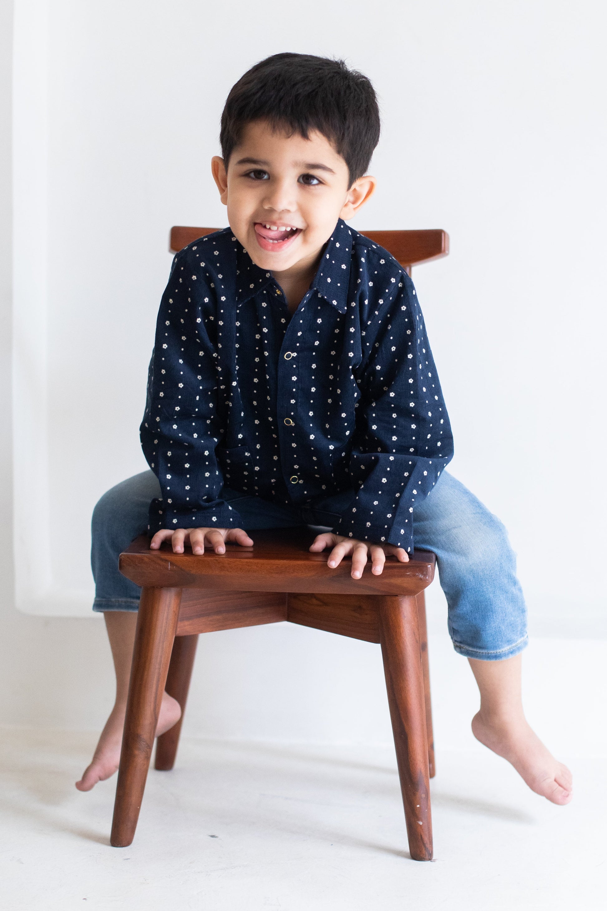 Child sitting on a wooden stool wearing a navy blue shirt with white patterns and light blue jeans.