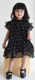 Young girl wearing a black floral dress standing on a white surface with a plain background