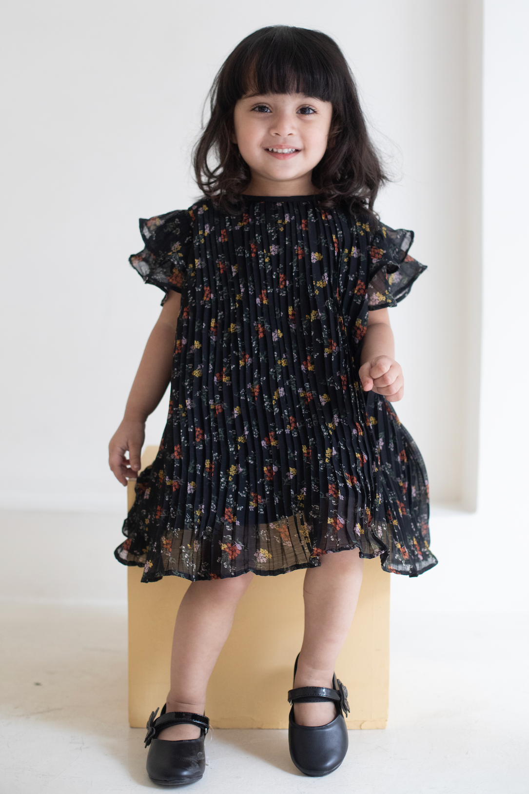 Young girl wearing a black floral dress standing on a white surface with a plain background