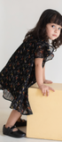 Young girl in a black floral dress standing on a beige block against a white background