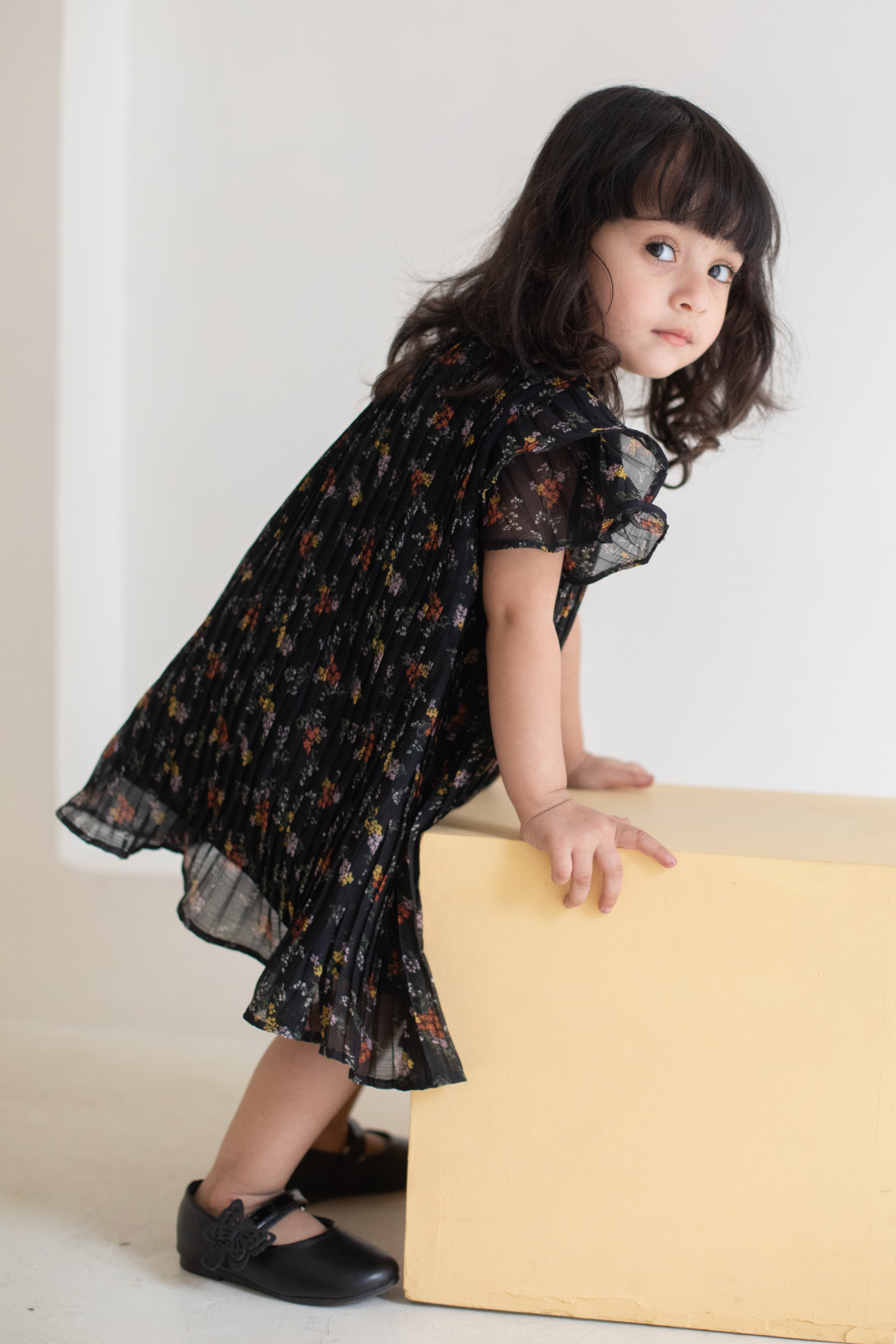 Young girl in a black floral dress standing on a beige block against a white background