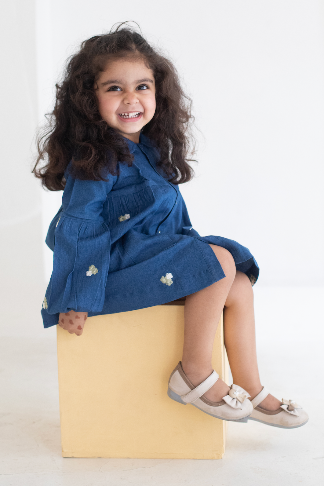 Child wearing a blue DRESS sitting on a yellow block against a white background