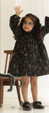 Young girl in a black floral dress standing in a softly lit room with a wooden chair and decorative garland.