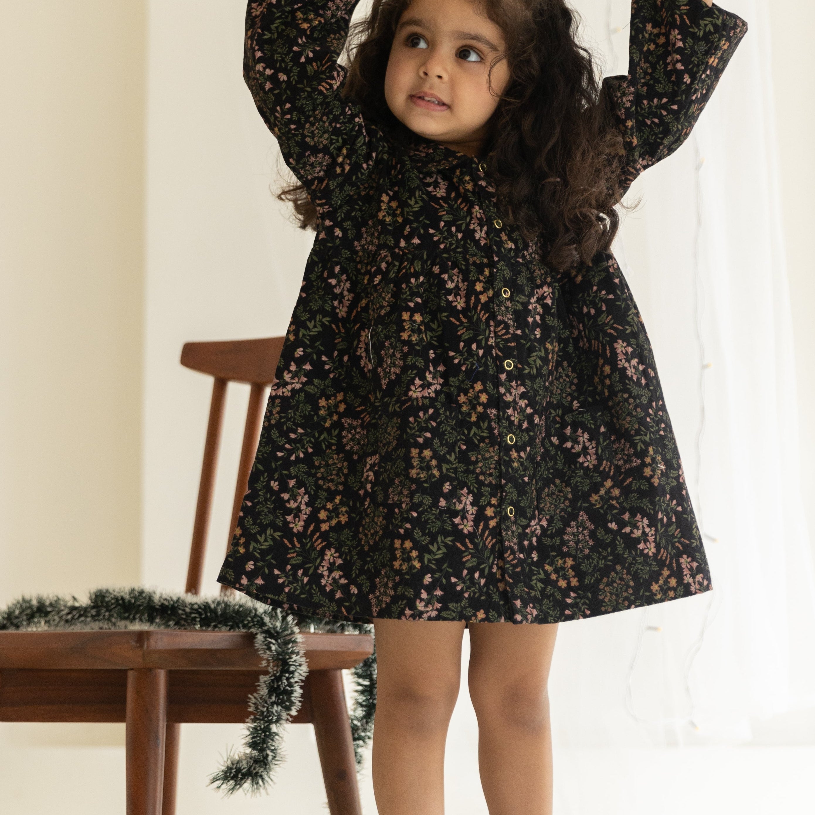 Young girl in a black floral dress standing in a softly lit room with a wooden chair and decorative garland.