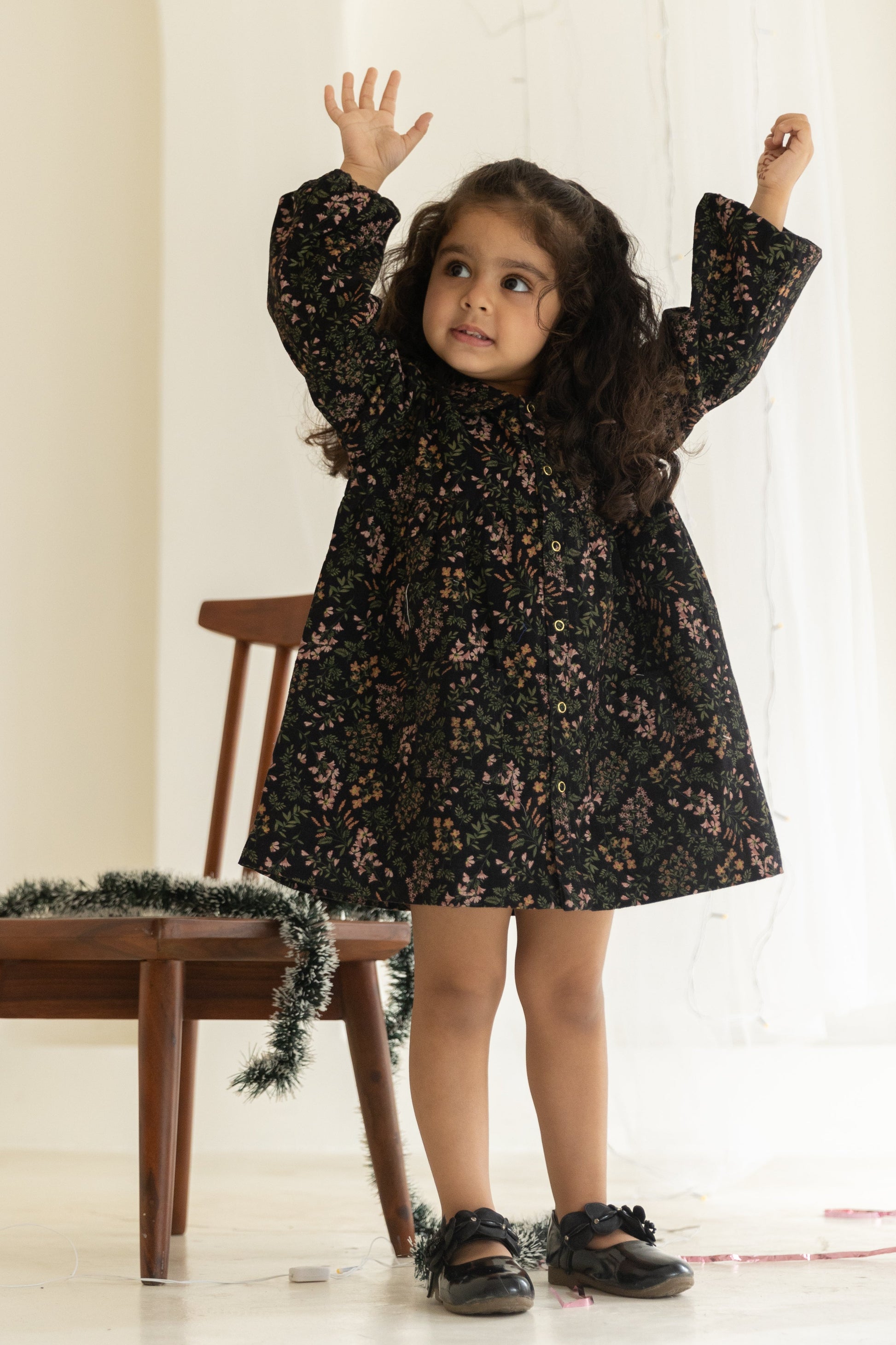 Young girl in a black floral dress standing in a softly lit room with a wooden chair and decorative garland.