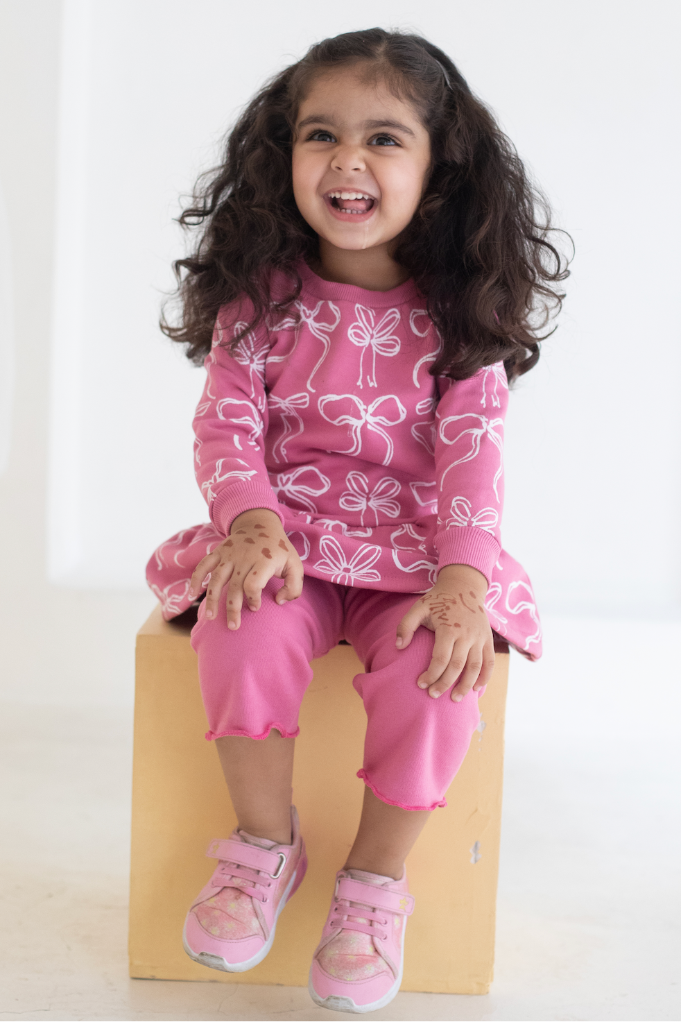 Child wearing an AUMORE KIDS pink outfit with bow patterns sitting on a wooden block against a white background