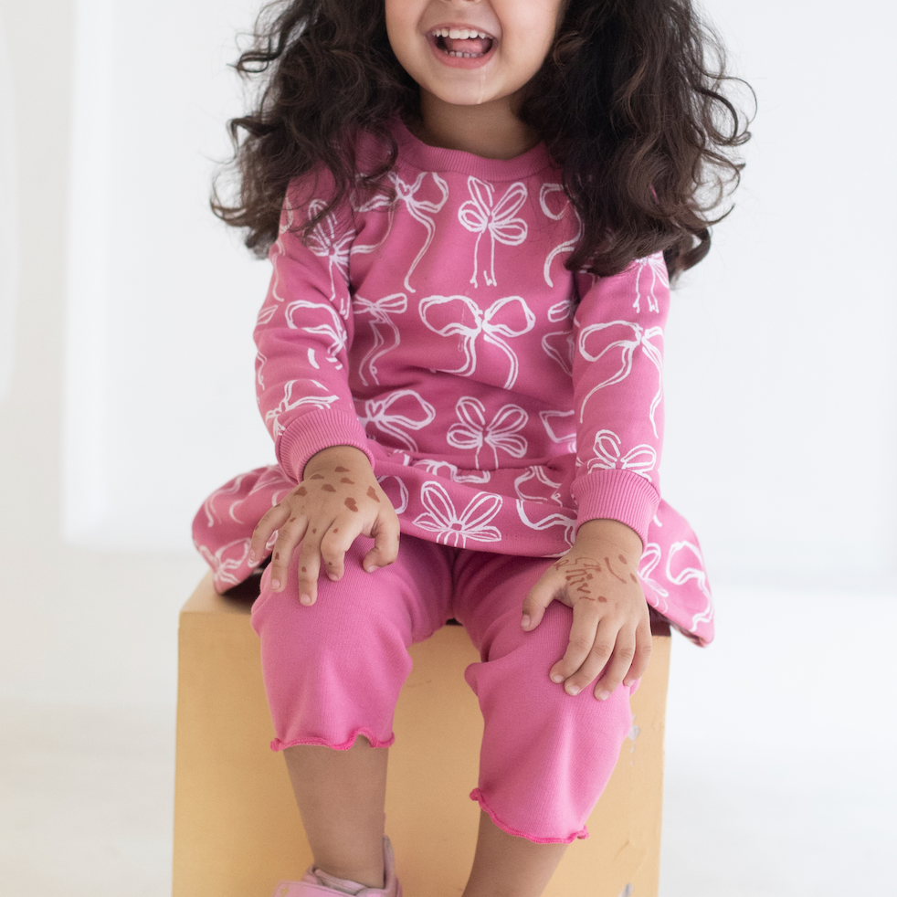 Child wearing an AUMORE KIDS pink outfit with bow patterns sitting on a wooden block against a white background