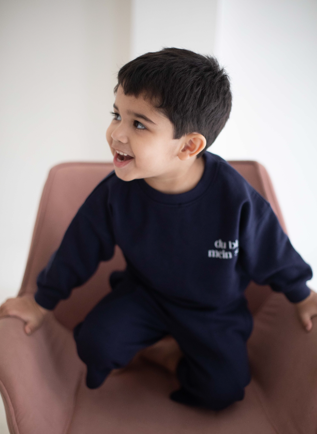 Child wearing a navy blue outfit with text, sitting on a pink chair.