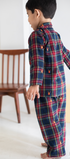 Child wearing a red and blue plaid outfit standing in a room with a wooden chair.