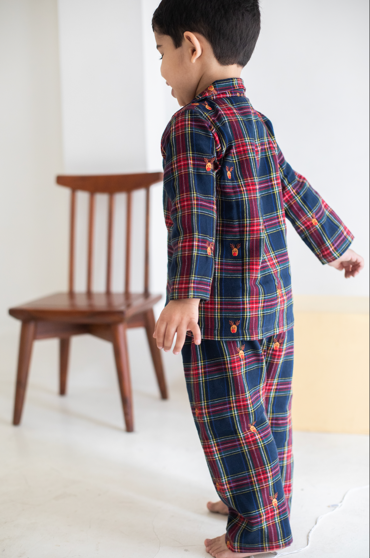 Child wearing a red and blue plaid outfit standing in a room with a wooden chair.