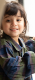 Child wearing a plaid coat standing by a window with curtains.