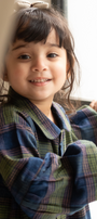 Child wearing a plaid coat standing by a window with curtains.