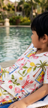 A young boy wearing a matching white short-sleeve camp shirt and shorts set with a colorful tropical lobster, lobster claw, and lobster tail print, lounging on a wicker lounge chair by a swimming pool.