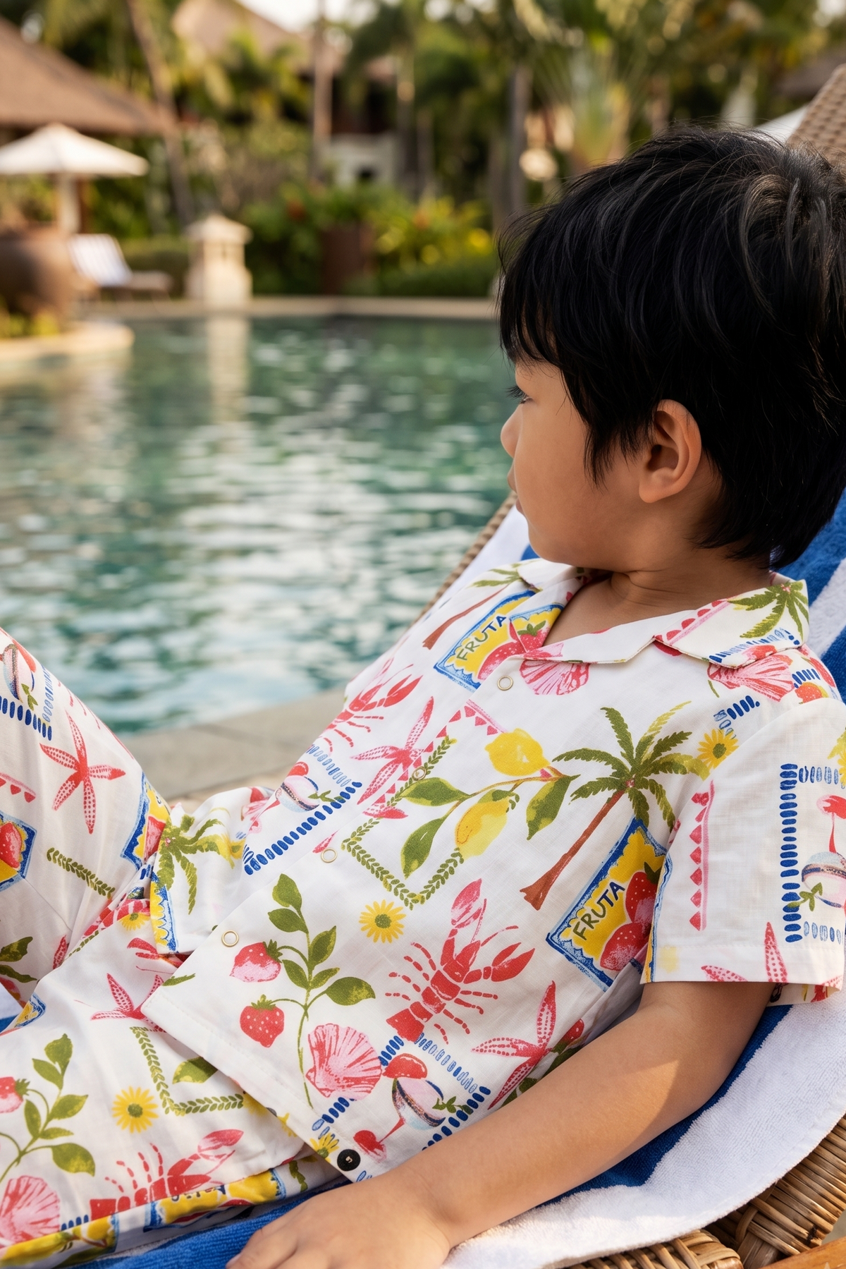 A young boy wearing a matching white short-sleeve camp shirt and shorts set with a colorful tropical lobster, lobster claw, and lobster tail print, lounging on a wicker lounge chair by a swimming pool.