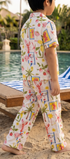 A toddler walking barefoot on a sandy beach poolside, wearing a short-sleeve matching pajama set with a colorful tropical print. The set features a white base with pink palm trees, blue stripes, and yellow sun motifs, with resort chairs and a swimming pool in the background.