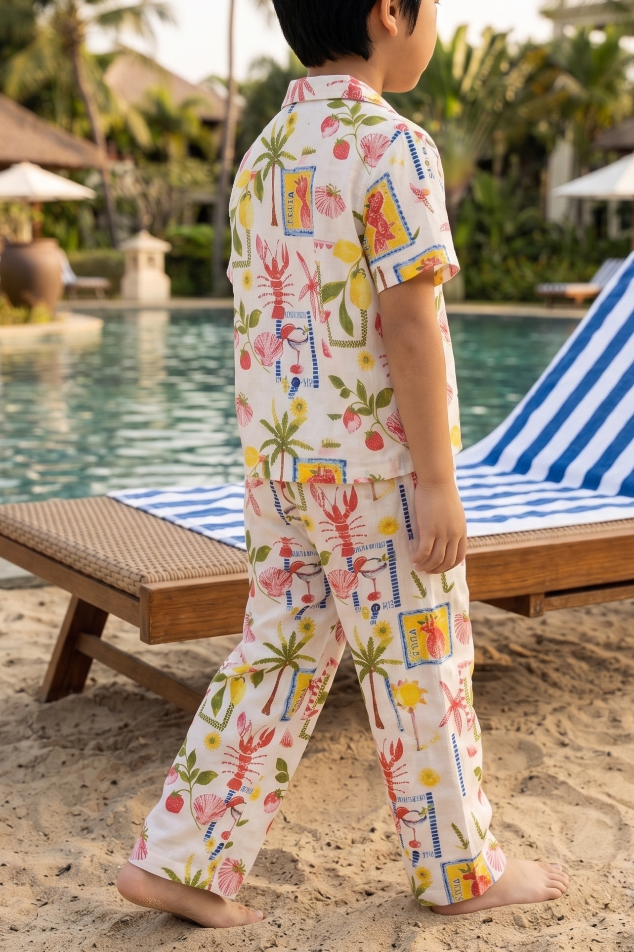 A toddler walking barefoot on a sandy beach poolside, wearing a short-sleeve matching pajama set with a colorful tropical print. The set features a white base with pink palm trees, blue stripes, and yellow sun motifs, with resort chairs and a swimming pool in the background.