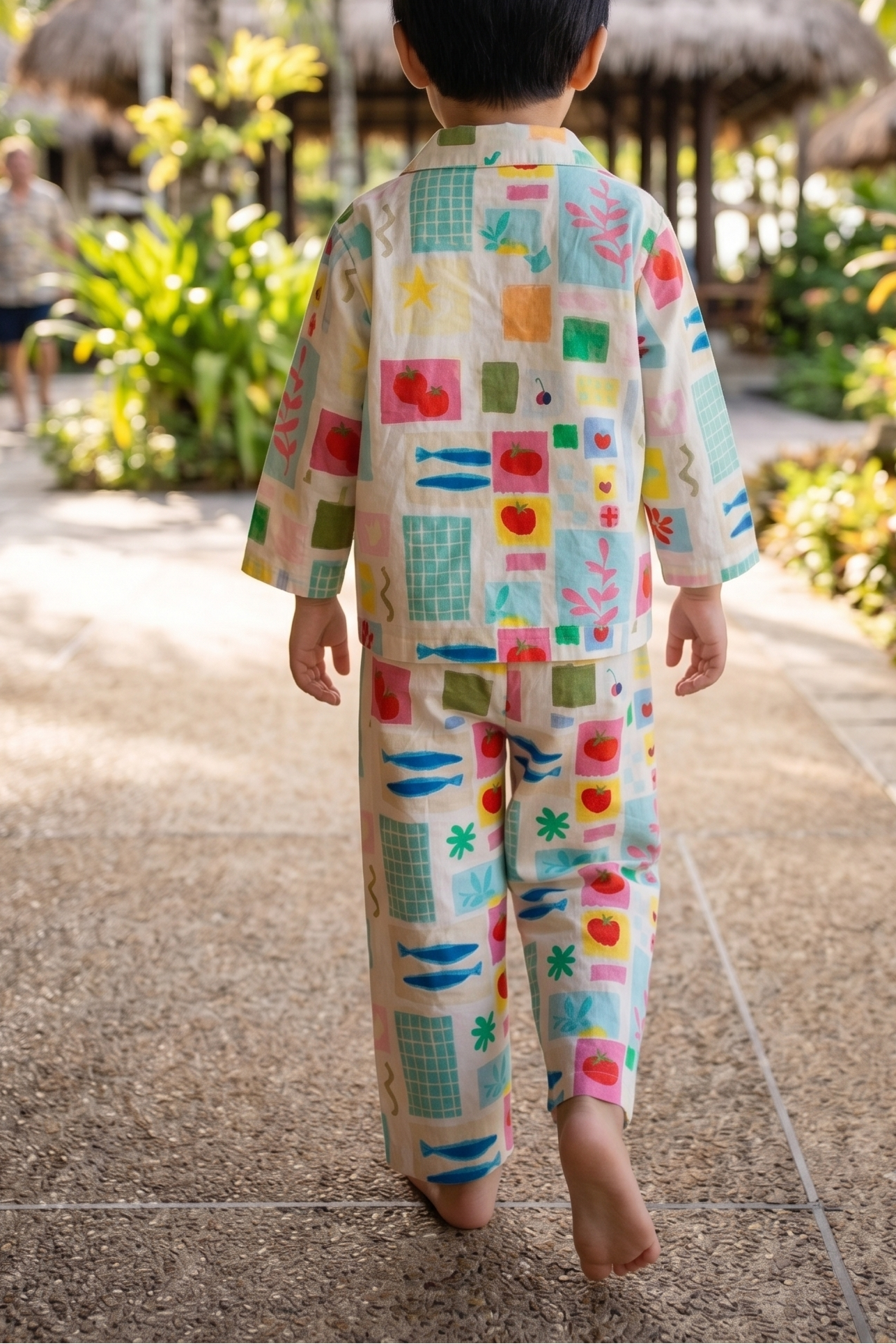 A child seen from behind walking barefoot on a tropical resort path, wearing a matching two-piece pajama set with a colorful patchwork print on a cream background.