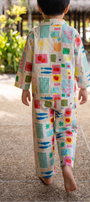 A child seen from behind walking barefoot on a tropical resort path, wearing a matching two-piece pajama set with a colorful patchwork print on a cream background.