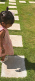 A toddler girl in a pink floral sleeveless dress walking on a grassy lawn with concrete stepping stones.