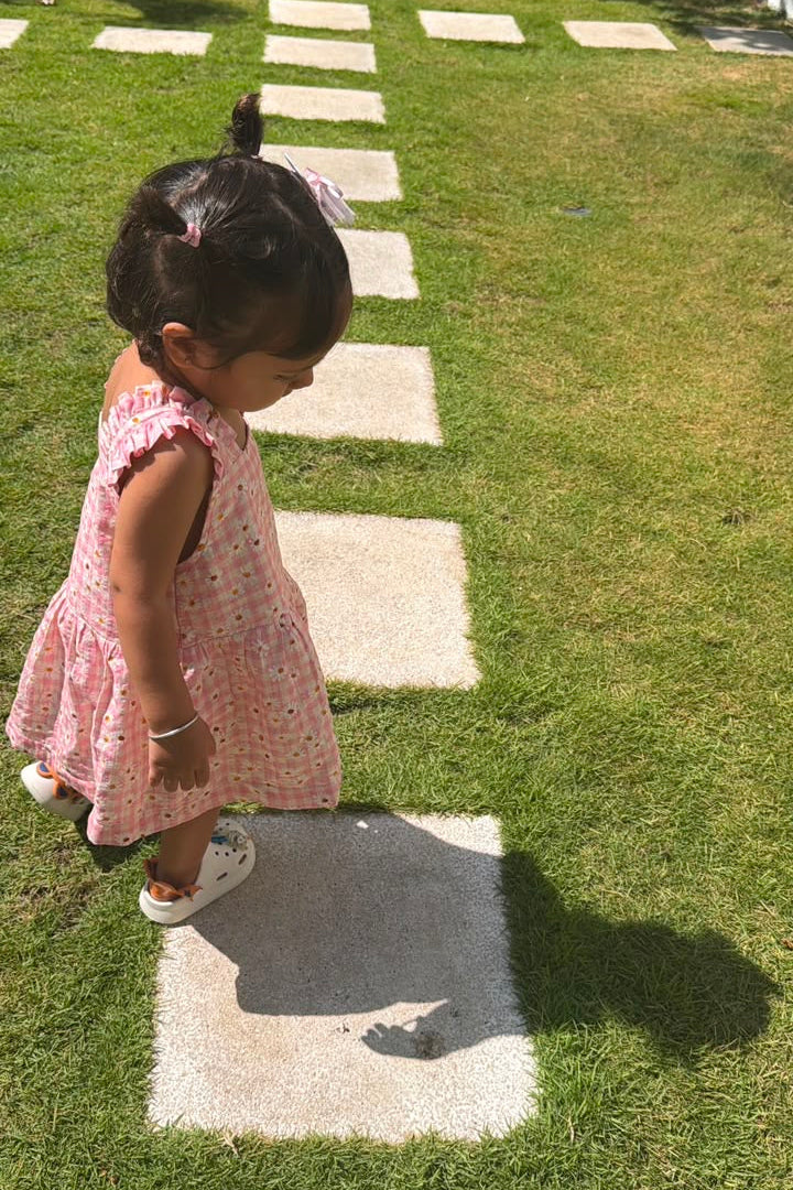 A toddler girl in a pink floral sleeveless dress walking on a grassy lawn with concrete stepping stones.