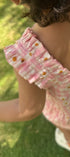 A toddler viewed from behind wearing a pink and white gingham ruffle-strap dress with small yellow and orange floral prints, standing on a green grassy lawn.