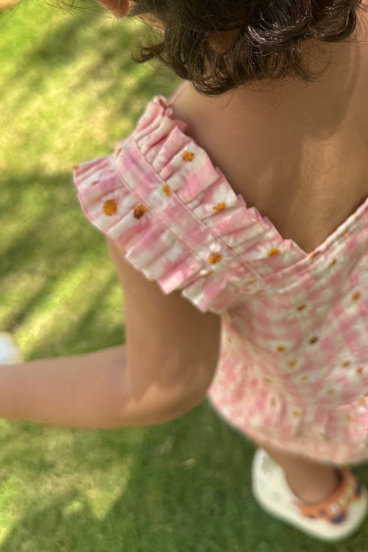 A toddler viewed from behind wearing a pink and white gingham ruffle-strap dress with small yellow and orange floral prints, standing on a green grassy lawn.