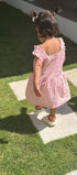 A toddler girl seen from behind wearing a pink floral off-the-shoulder dress and white sneakers, walking on a green lawn between concrete stepping stones.
