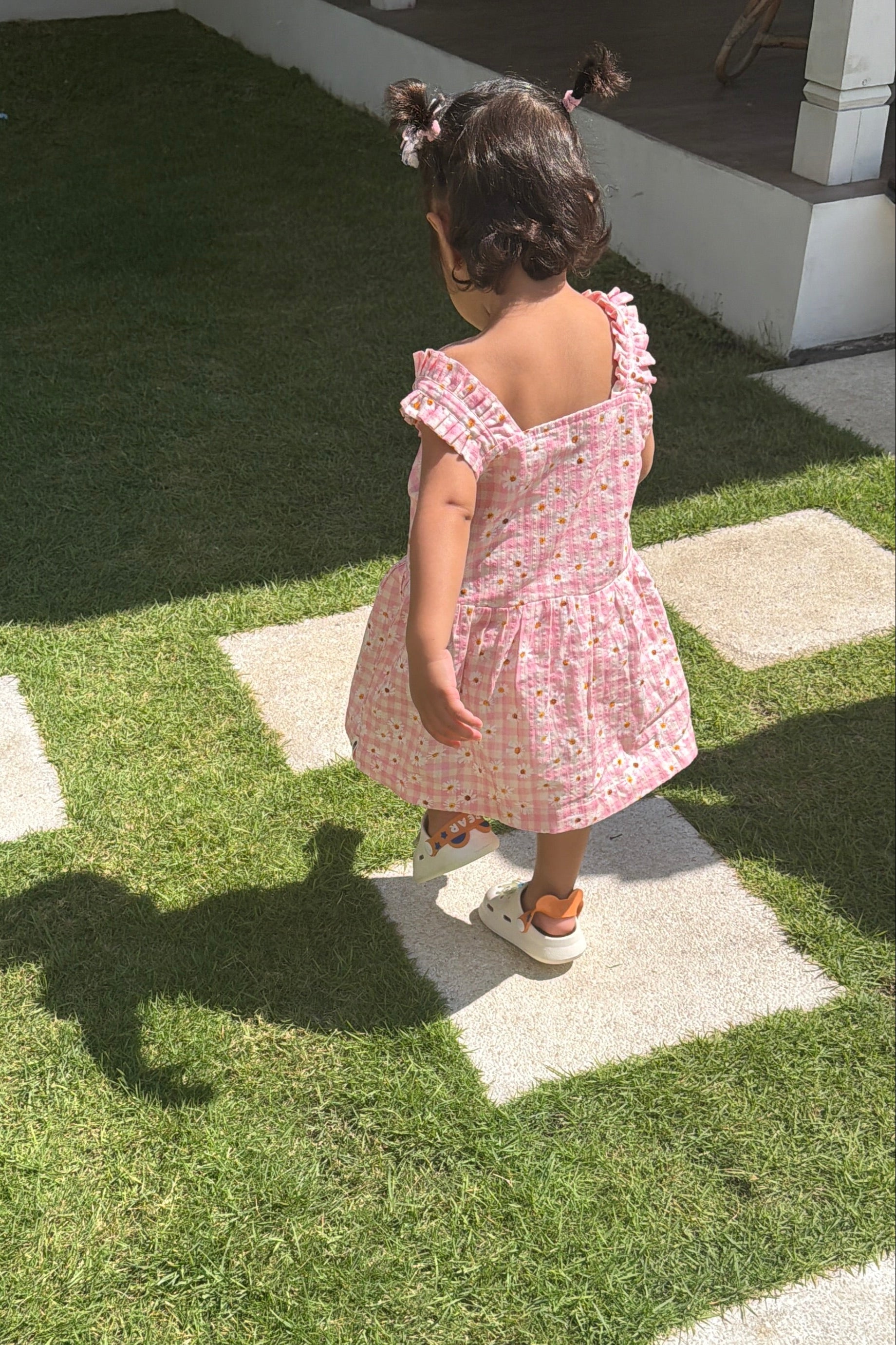 A toddler girl seen from behind wearing a pink floral off-the-shoulder dress and white sneakers, walking on a green lawn between concrete stepping stones.