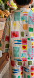 A child viewed from behind wearing a matching two-piece pajama set with a colorful patchwork print of fruit, fish, stars, and geometric shapes, standing in a tropical resort setting.